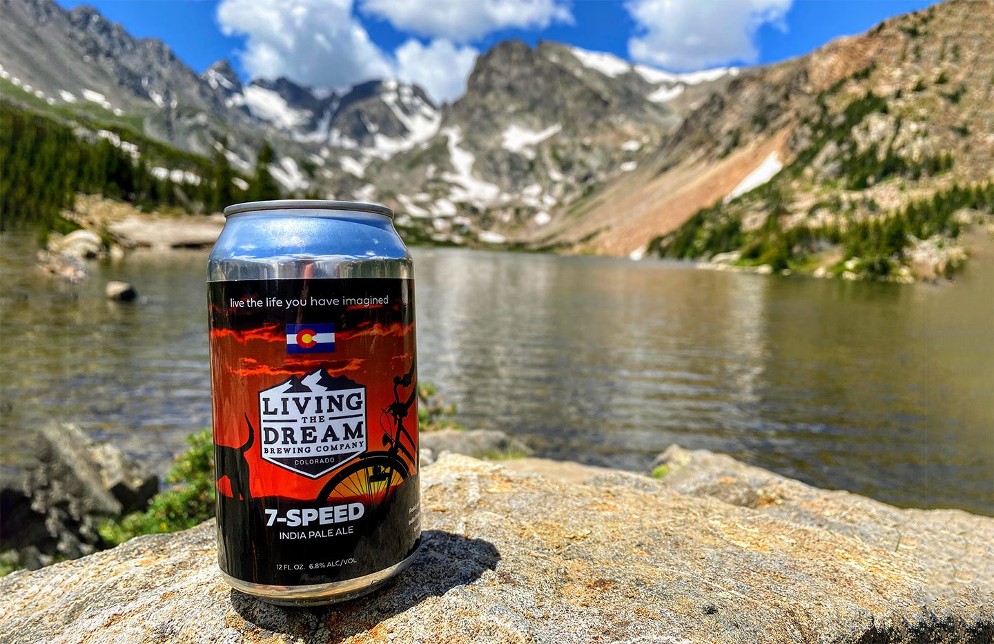 Can of 7-Speed IPA beer sitting on rock near edge of water with Colorado mountains in the back